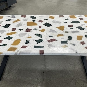 Peut inclure: Une table rectangulaire dotée d'une surface blanche incrustée d'un motif terrazzo multicolore composé de formes géométriques dorées, vert foncé et bordeaux. La table est soutenue par deux pieds en métal noir en forme de U à chaque extrémité.