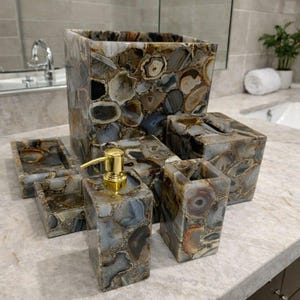 Puede incluir: Un juego de baño que incluye un dispensador de jabón con bomba dorada, una caja de pañuelos, un porta cepillos de dientes, una bandeja y un cesto de basura. El juego está hecho de piedra con un patrón de mosaico en tonos grises, marrones y crema.