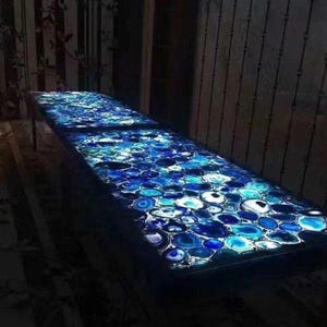 May include: A long, rectangular table with a surface made of blue and white agate stones. The stones are arranged in a mosaic pattern, and the table is illuminated from within, creating a glowing effect. The table is set in a dimly lit room.