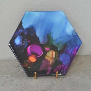 May include: Hexagon-shaped abstract art piece with a glossy finish. The artwork features a blend of vibrant colors, including blues, purples, pinks, and oranges, creating a dynamic and fluid design. The piece is displayed on a gold-colored stand.