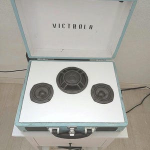 May include: A light blue suitcase-style Victrola record player with the lid open. The interior features a white panel mounted with three black circular speakers of varying sizes. The device includes a black carrying handle and metal latches.