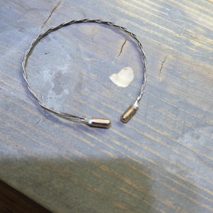 May include: A silver-toned, twisted wire bracelet with gold-colored end caps. The bracelet is open, with the ends not connected. The bracelet is on a weathered, gray wooden surface.