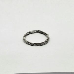 May include: A silver ring crafted from tightly wound guitar strings. The ring has a textured, metallic appearance and a circular shape. The design features multiple strands of wire, creating a unique and artistic piece of jewelry.