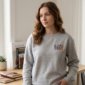 May include: A light gray sweatshirt with a small embroidered design of colorful jars with flowers. The sweatshirt is crew neck and has long sleeves. The person in the image is wearing a gold necklace.
