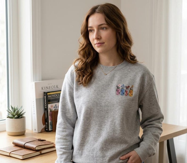 May include: A light gray sweatshirt with a small embroidered design of colorful jars with flowers. The sweatshirt is crew neck and has long sleeves. The person in the image is wearing a gold necklace.