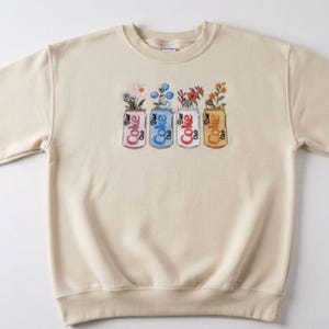 May include: Cream-colored sweatshirt with a crew neck and long sleeves. The front features an embroidered design of four Coke cans, each holding a different arrangement of flowers. The cans are in pink, blue, white, and gold.
