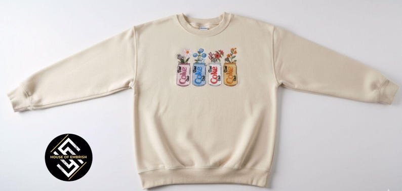May include: Cream-colored sweatshirt with a crew neck and long sleeves. The front features an embroidered design of four Coke cans, each holding a different arrangement of flowers. The cans are in pink, blue, white, and gold.