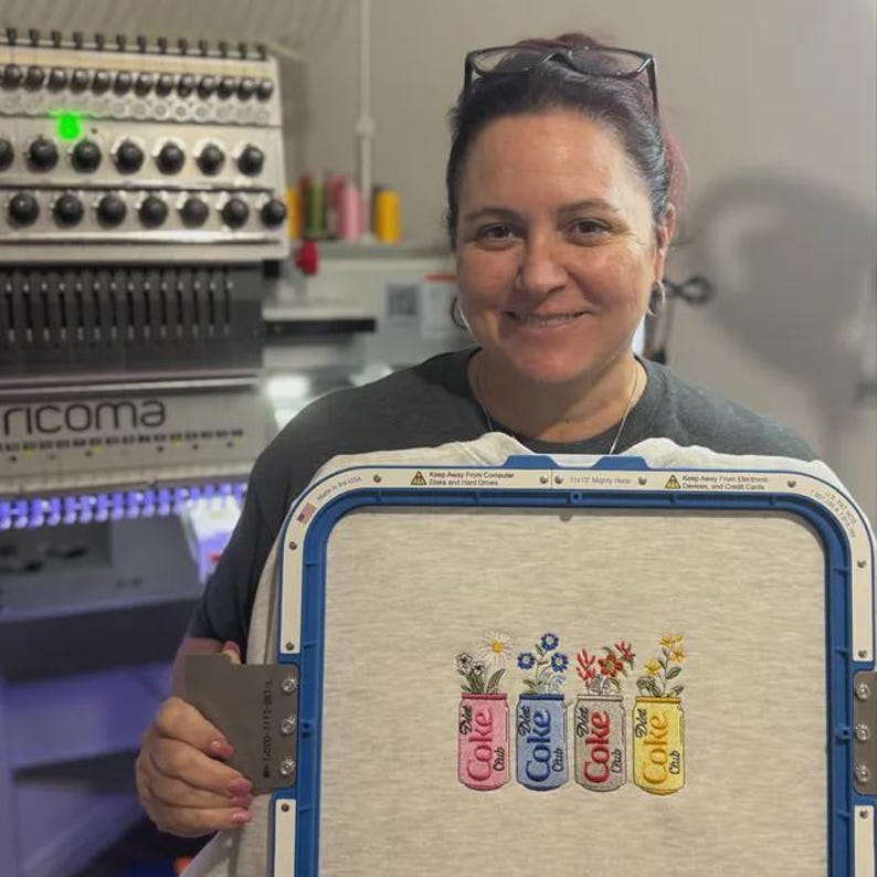 May include: A woman holds a light gray fabric in an embroidery hoop. The fabric features an embroidered design of four Diet Coke cans, each with a different color of flowers. The cans are red, blue, red, and yellow.