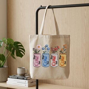 May include: Beige tote bag featuring four Diet Coke cans, each holding different flowers. The cans are pink, blue, red, and yellow, with the words "Diet Coke Club" printed on them. The bag hangs on a black metal bar.