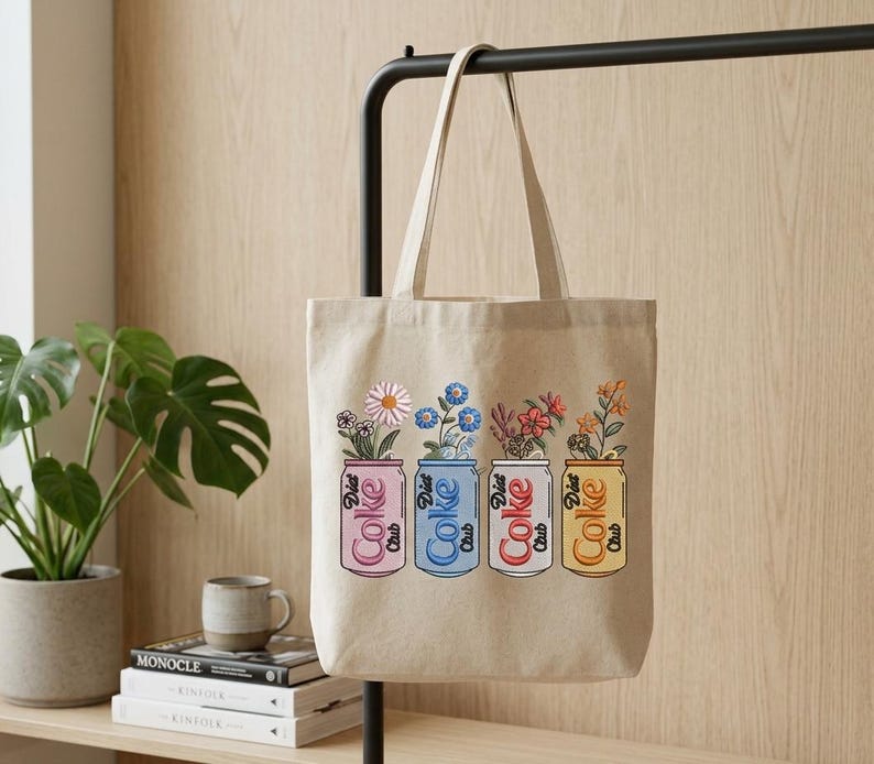 May include: Beige tote bag featuring four Diet Coke cans, each holding different flowers. The cans are pink, blue, red, and yellow, with the words "Diet Coke Club" printed on them. The bag hangs on a black metal bar.