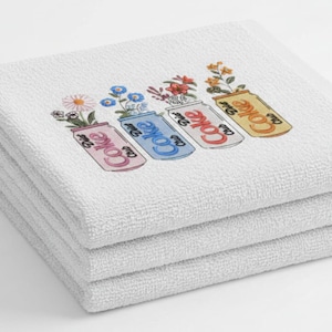 May include: A stack of four white, fluffy towels. Each towel is embroidered with a colorful design of a Coke can filled with flowers. The cans are pink, blue, red, and yellow. The text "Coke" is visible on each can.