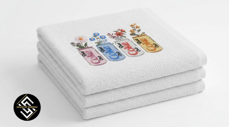 May include: A stack of four white, fluffy towels. Each towel is embroidered with a colorful design of a Coke can filled with flowers. The cans are pink, blue, red, and yellow. The text "Coke" is visible on each can.