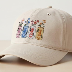 May include: A beige baseball cap featuring a colorful graphic of four Coke cans, each filled with different flowers. The cans are pink, blue, red, and yellow, with the word "Coke" printed on them. The cap has a curved brim.
