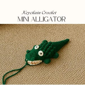 May include: A handmade green crochet alligator keychain with white eyes and a smiling mouth. The keychain is attached to a green string. Text above the alligator reads "Keychain Crochet MINI ALLIGATOR".