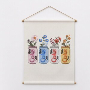 May include: A wall hanging featuring four Diet Coke cans, each holding a different arrangement of flowers. The cans are pink, blue, red, and yellow, with the words "Diet Coke Club" printed on them. The artwork is displayed on a cream-colored fabric.