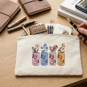 May include: A cream-colored canvas zippered pouch with an embroidered design of four Diet Coke cans, each holding different flowers. The cans are in pink, blue, red, and yellow, with the words "Diet Coke Club" on them. Other stationery items are visible.