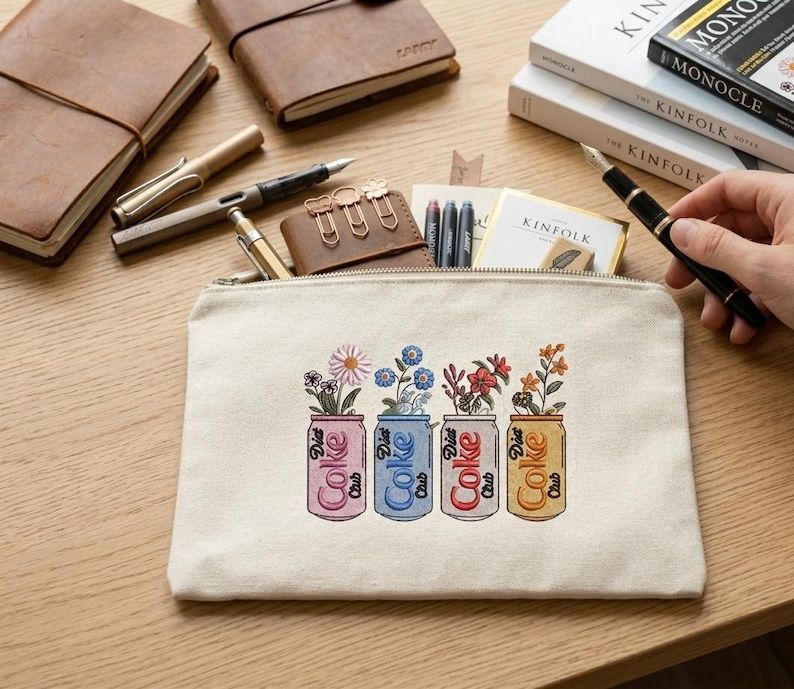 May include: A cream-colored canvas zippered pouch with an embroidered design of four Diet Coke cans, each holding different flowers. The cans are in pink, blue, red, and yellow, with the words "Diet Coke Club" on them. Other stationery items are visible.