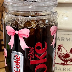 May include: A clear glass jar filled with a dark liquid, ice, and decorated with pink bows and "Diet Coke" branding. The jar has a clear lid and is next to a ceramic container with a rooster illustration.