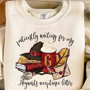 Puede incluir: Sudadera color crema con un diseño que incluye un sombrero de bruja, libros, una taza roja con crema batida y el texto "patiently waiting for my Hogwarts acceptance letter."