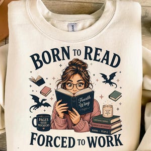 Puede incluir: Una sudadera de cuello redondo color crema con un gráfico de una persona leyendo un libro titulado Fourth Wing. El texto dice Born to Read Forced to Work. Incluye elementos como dragones voladores, pilas de libros, una vela y una taza.
