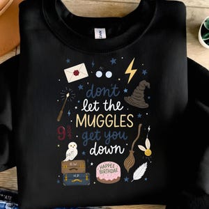May include: Black sweatshirt with a whimsical design featuring a wand, owl, and the text "don't let the Muggles get you down." Other graphics include a lightning bolt, sorting hat, and a cake that says "Happee Birthdae."