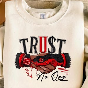 以下が含まれることがあります： 黒と赤で「TRUST」の文字がプリントされたクリーム色のスウェットシャツ。 「U」にはドル記号が組み込まれています。 下には、握手の周りにヘビが巻き付いたグラフィックと「No One」の文字があります。