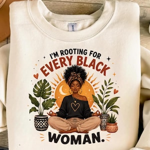 May include: A cream-colored crewneck sweatshirt featuring a graphic print of a person with dark skin and curly hair sitting in a lotus position. The text reads I'M ROOTING FOR EVERY BLACK WOMAN, surrounded by potted plants and a sun illustration.