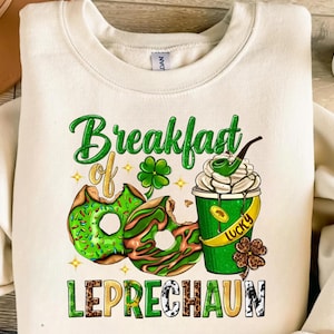 Puede incluir: Sudadera de color crema con un diseño del Día de San Patricio. El diseño presenta rosquillas verdes, una taza de café con crema batida y el texto "Breakfast of Leprechaun". El diseño también incluye tréboles y la palabra "Lucky".