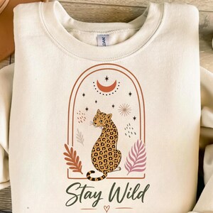 Peut inclure: Sweat-shirt crème avec une illustration de léopard dans une arche marron, un croissant de lune et des étoiles. Les mots "Stay Wild" sont écrits en dessous dans une police script. Le sweat-shirt a un col rond.
