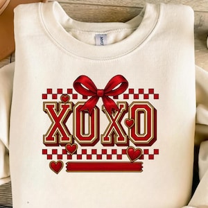 Puede incluir: Sudadera color crema con un diseño rojo y dorado. El diseño presenta las letras "XOXO" con un lazo rojo, corazones y un patrón de cuadros. Un gráfico temático del Día de San Valentín.
