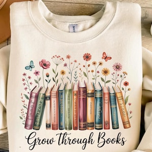 May include: Cream-coloured sweatshirt featuring a graphic of books, flowers, and butterflies. The phrase "Grow Through Books" is printed in a script font. The sweatshirt has a crew neck. The design is colourful.