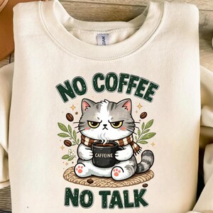 May include: A cream-colored crewneck sweatshirt featuring a graphic of a grumpy gray and white cat wearing a striped scarf. The cat holds a black mug labeled CAFFEINE. The text NO COFFEE NO TALK is printed in dark green above and below the cat.