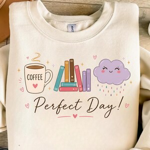 May include: A cream-colored crewneck sweatshirt featuring a graphic print of a coffee mug labeled COFFEE, a stack of colorful books, and a smiling purple rain cloud. Below the illustrations, the text Perfect Day! is printed in black script.