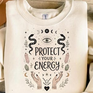 May include: Cream-coloured sweatshirt with a graphic design featuring the text "PROTECT YOUR ENERGY". The design includes snakes, crystals, moons, stars, an eye, and hands. The sweatshirt is a crew neck style.
