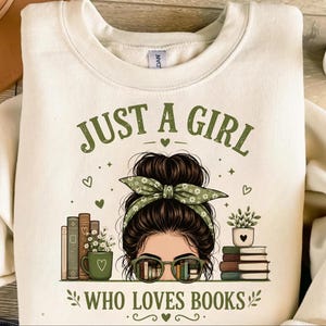 May include: Cream sweatshirt with the text "JUST A GIRL WHO LOVES BOOKS" in green. The graphic features a woman with glasses, a green headband, and books. The design includes hearts and floral accents.