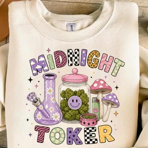 Puede incluir: Una sudadera de cuello redondo color crema con un estampado gráfico que incluye el texto MIDNIGHT TOKER. El diseño muestra una pipa de agua de vidrio morada, un frasco transparente con cogollos verdes, dos setas, un encendedor y un pequeño recipiente rosa.