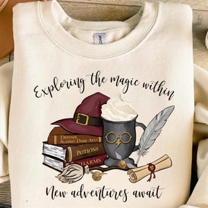 May include: Cream-colored sweatshirt with a graphic featuring a wizard hat, books, a coffee cup with glasses, a feather, and a scroll. Text reads "Exploring the magic within" and "New adventures await."