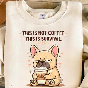 May include: Cream-colored sweatshirt with the text "THIS IS NOT COFFEE. THIS IS SURVIVAL." features a cartoon French Bulldog holding a coffee cup. The dog has a grumpy expression and is surrounded by small hearts and stars.