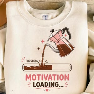 May include: A cream-coloured sweatshirt featuring a coffee-themed design. A pink and brown coffee maker pours coffee into a progress bar that reads "Motivation Loading...". The word "Progress" is above the bar. A small succulent plant is visible in the upper right corner.
