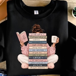 Könnte beinhalten: Ein schwarzes Sweatshirt mit Rundhalsausschnitt und einer Grafik einer Person mit braunem Dutt, die im Schneidersitz sitzt. Die Person trägt einen rosa Pullover und hält ein offenes Buch mit dem Titel one more chapter sowie eine Tasse.