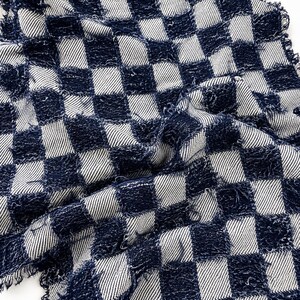 May include: A fabric textile featuring a dark navy blue and white checkered pattern. The material has a textured, frayed appearance with visible loose threads along the edges of the squares, creating a distressed and woven look throughout the cloth.
