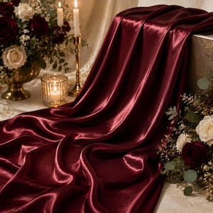 Può includere: Un tessuto di raso color bordeaux intenso drappeggiato in morbide pieghe, caratterizzato da una superficie liscia e riflettente. Il tessuto è posizionato vicino a un vaso dorato con rose bianche e rosso scuro, un portacandele acceso e una candela.