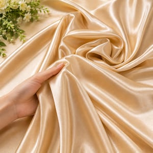 May include: A piece of champagne-coloured satin fabric with a smooth, reflective surface and deep, flowing folds. The material has a high-sheen finish that catches the light, creating bright highlights and soft shadows across the draped textile.