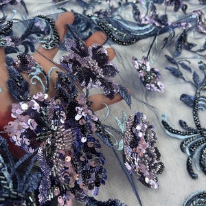 Puede incluir: Primer plano de una tela de tul azul marino con intrincados bordados florales. El diseño presenta racimos de lentejuelas lavanda y plateadas, cuentas y detalles bordados. La tela es ideal para ropa de noche o de ocasión especial.