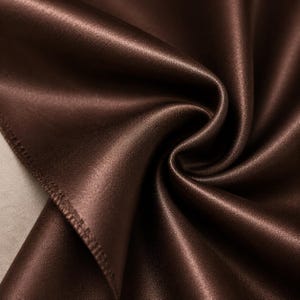 Könnte beinhalten: Ein dunkelbrauner Stoff mit einer glatten, glänzenden und satinartigen Oberfläche. Das Material ist in weichen, fließenden Falten drapiert, die tiefe Schatten werfen und die reflektierende Beschaffenheit sowie den satten Schokoladenbraunton betonen.