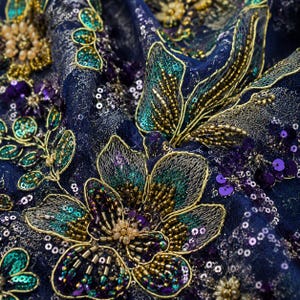 May include: A deep blue fabric featuring intricate floral embroidery with metallic gold thread, iridescent teal sequins, and purple beads. The design includes detailed petal outlines and clusters of small, shimmering beads arranged in floral patterns.