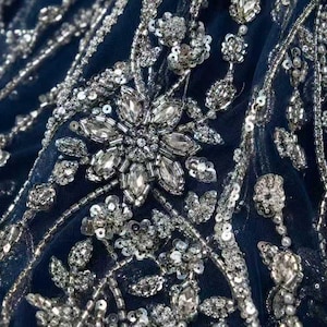 Pode incluir: Close-up de tecido azul marinho adornado com lantejoulas, contas e cristais prateados. O design apresenta um grande motivo floral central e pequenos grupos florais, criando uma textura luxuosa e detalhada.