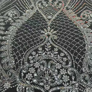 May include: Detailed shot of black fabric adorned with silver beads and sequins. The design showcases floral and geometric motifs, forming an opulent and elaborate texture. The shimmering embellishments give the textile a touch of sophistication.