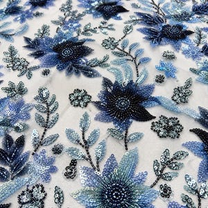 Puede incluir: Primer plano de una tela con diseño floral. La tela presenta flores bordadas en tonos azules, con intrincados detalles de cuentas y lentejuelas. Las flores están dispuestas sobre un fondo blanco, creando una superficie texturizada y decorativa.