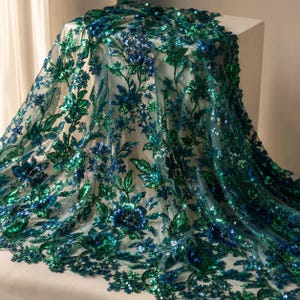 May include: A sheer fabric featuring an intricate floral pattern embroidered with vibrant emerald green and deep royal blue sequins. The design showcases detailed leaves and blossoms, creating a dense, shimmering texture across the entire surface.